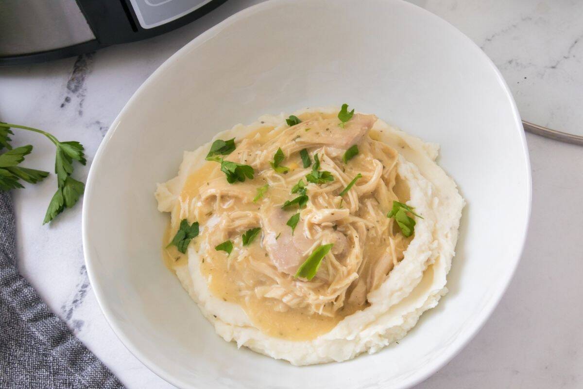A bowl of mashed potatoes topped with shredded chicken in creamy gravy, garnished with chopped parsley.