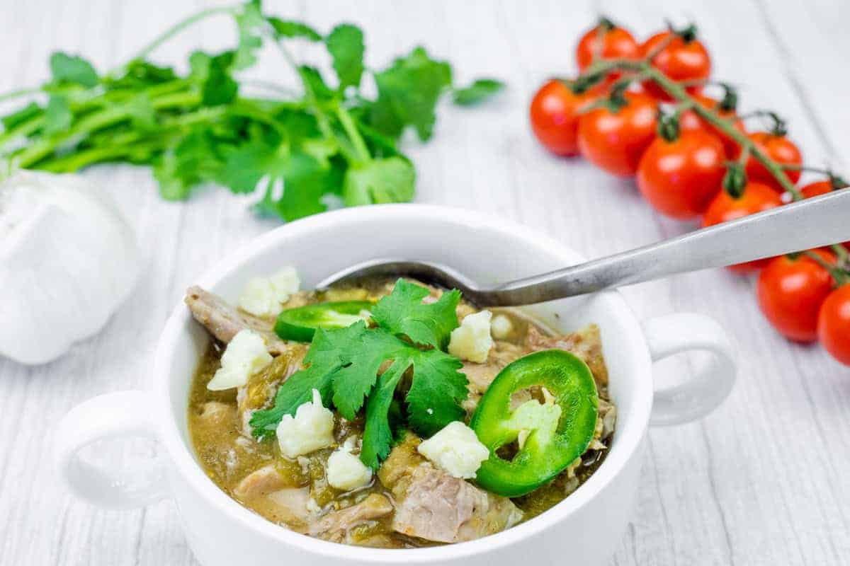 Close-up of a bowl of slow cooker pork chile verde served with white rice, garnished with fresh cilantro, sliced jalapeños, radish slices, and a wedge of lime.
