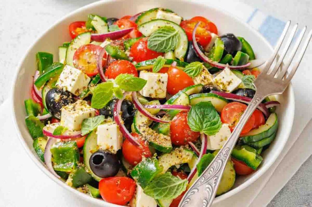 A bowl of Greek salad with cherry tomatoes, cucumber, red onion, green pepper, black olives, feta cheese, and mint leaves, served with a fork.