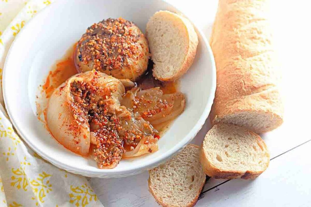 A white bowl with spicy stuffed onions in sauce, served with sliced baguette bread on the side.