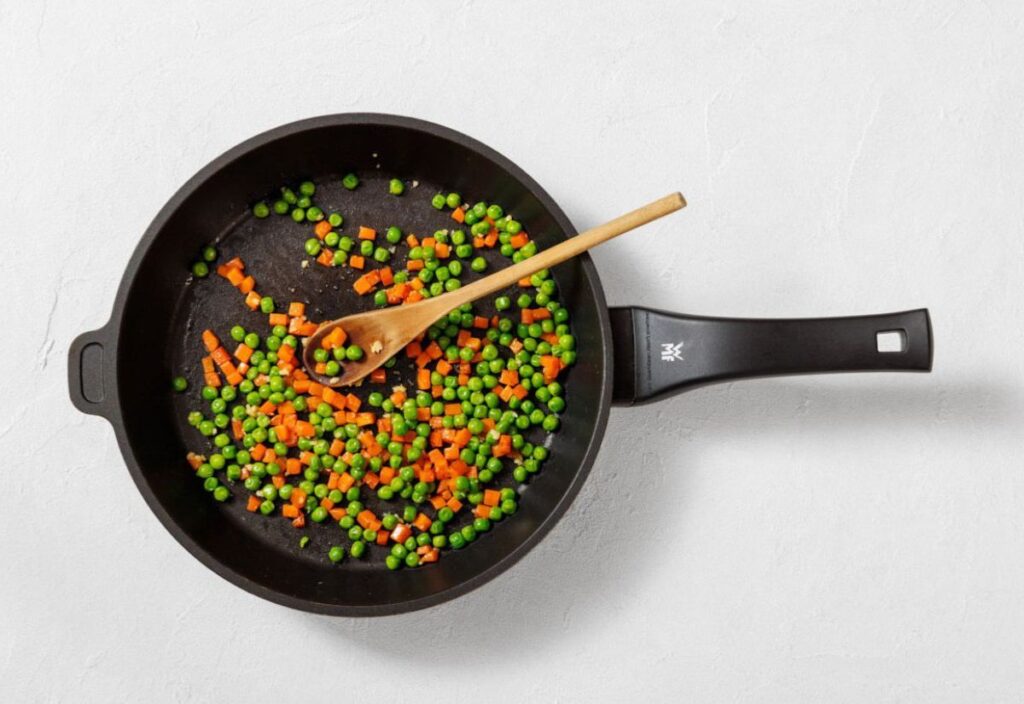 A skillet with diced carrots and peas sautéing on medium heat, being stirred with a wooden spoon.