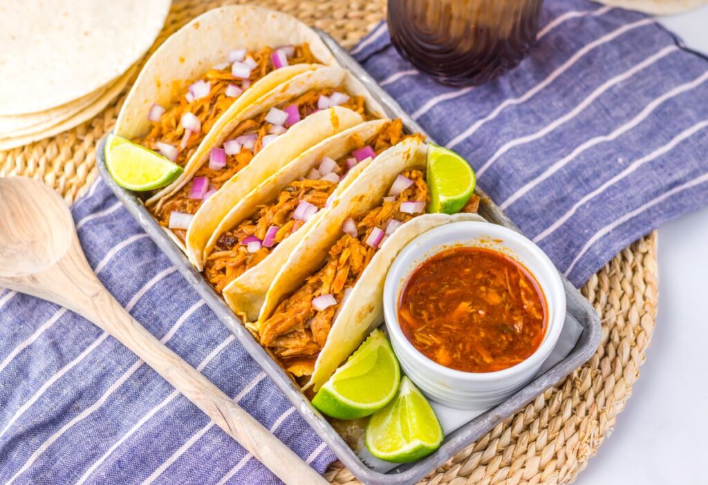 Tray of shredded pork tacos with diced onions and lime wedges, accompanied by a bowl of dipping sauce.