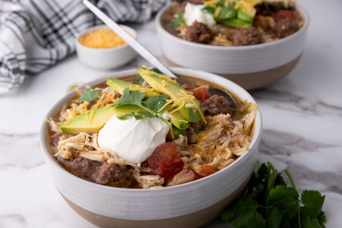 Two bowls of chili topped with avocado slices, sour cream, shredded cheese, and cilantro. Sliced lime and cheese bowl in the background on a marble surface.
