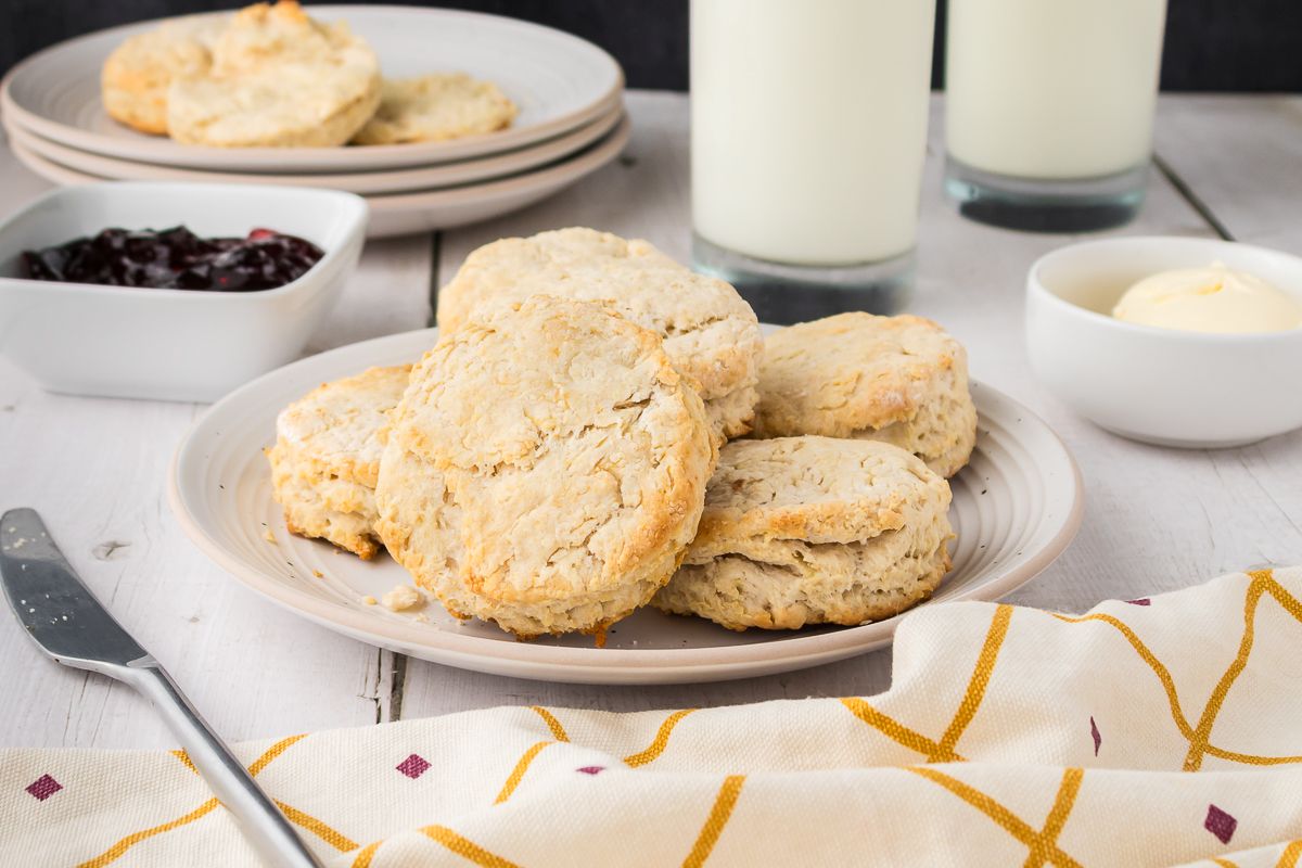 A plate of biscuits is on a table with a knife, a bowl of butter, a bowl of jam, two glasses of milk, and a yellow-patterned napkin.
