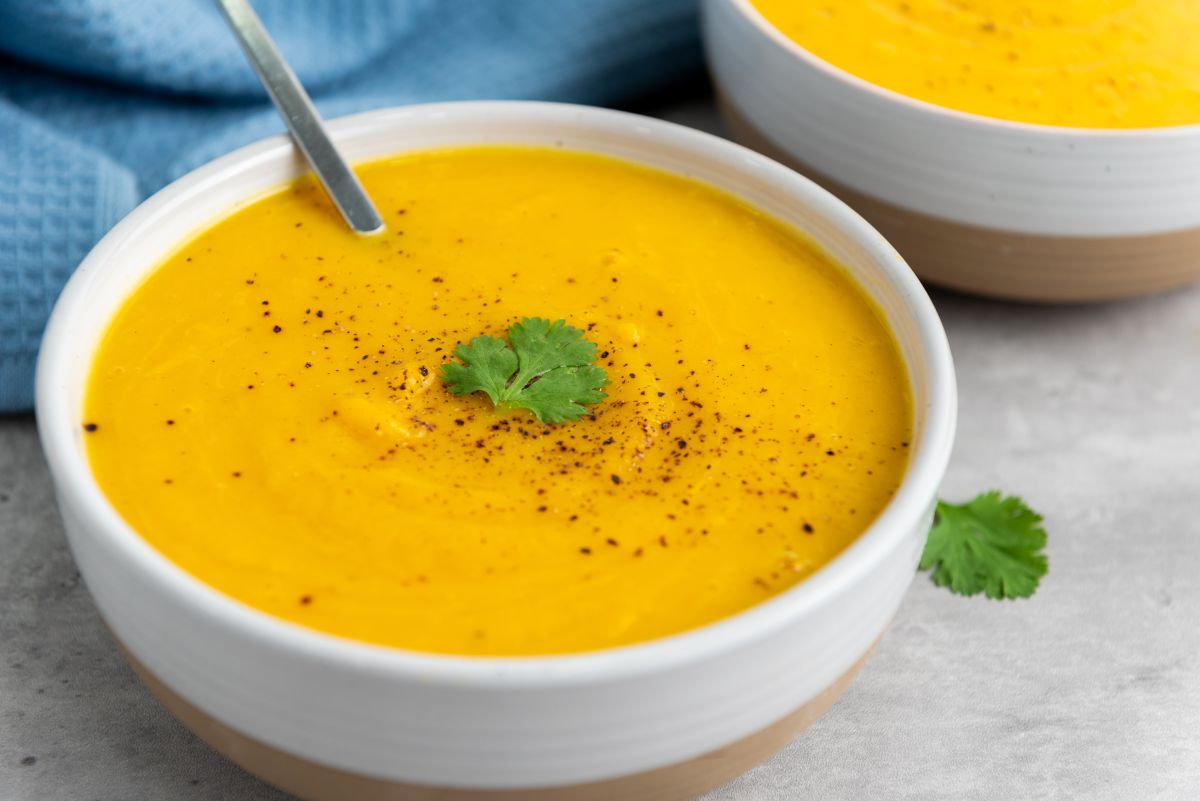A bowl of creamy orange soup garnished with black pepper and a cilantro leaf, with a spoon resting inside the bowl. Another identical bowl is partially visible in the background.