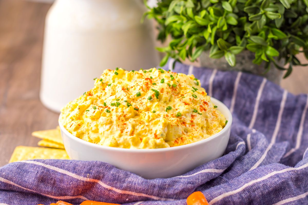 A bowl of deviled egg dip garnished with chives and paprika, surrounded by baby carrots on a striped cloth.