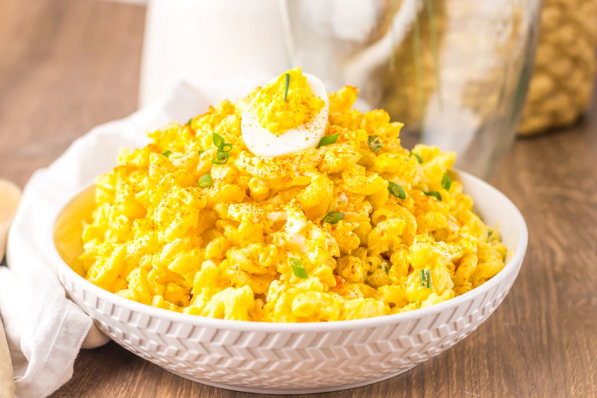A bowl of creamy macaroni salad topped with sliced boiled eggs, chopped green onions, and a sprinkle of paprika, placed on a wooden surface.