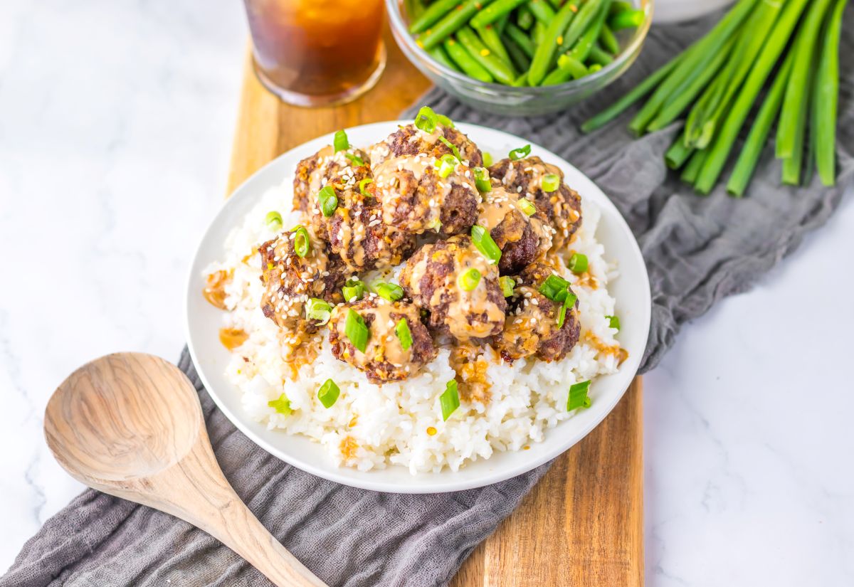 A plate of white rice topped with saucy meatballs, garnished with chopped green onions, sits on a wooden board with a wooden spoon and a drink in the background.