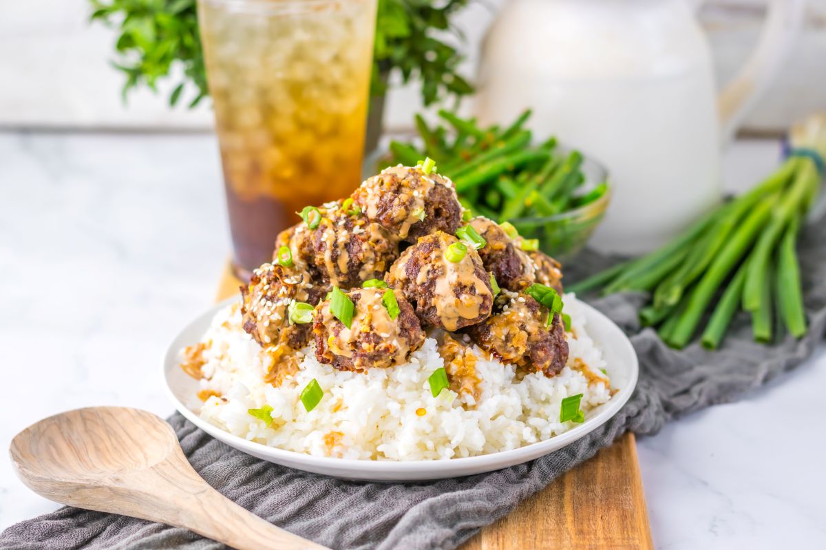 A plate of white rice topped with saucy meatballs, garnished with chopped green onions, sits on a wooden board with a wooden spoon and a drink in the background.