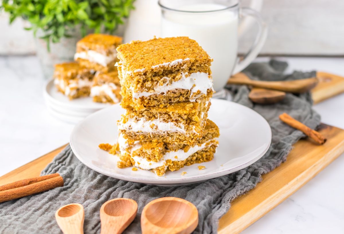 Three oatmeal cream pies are stacked on a white plate, filled with white cream, with a glass of milk, cinnamon sticks, and wooden spoons nearby on a wooden tray.