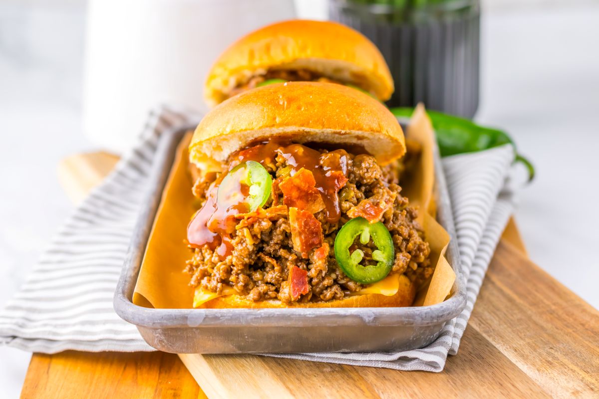 Two sloppy joe sandwiches topped with sliced jalapeños and sauce are served on a metal tray lined with parchment paper, with a striped napkin underneath.