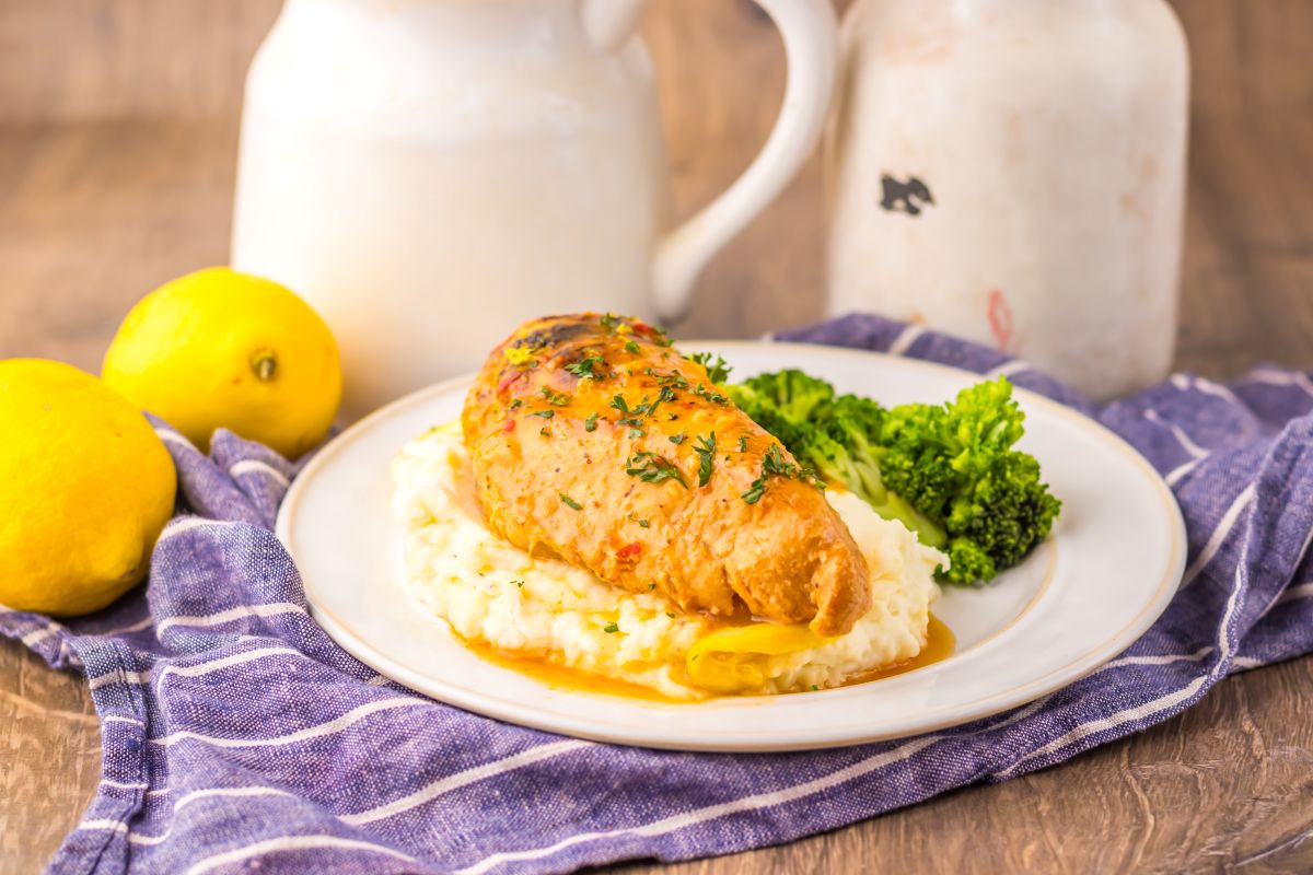 A stuffed chicken breast topped with herbs is served on mashed potatoes with a side of steamed broccoli on a white plate.
