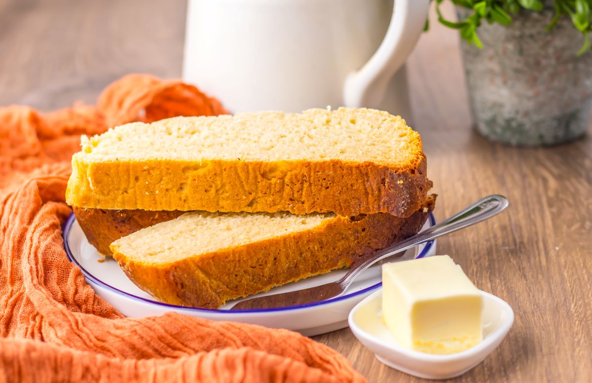 Two slices of bread on a plate with a butter knife, next to a small dish of butter, an orange cloth, and a white pitcher in the background.