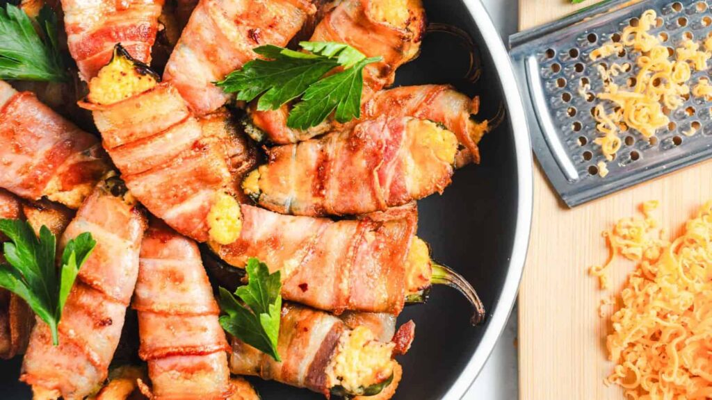 Plate of bacon-wrapped peppers filled with cheese, garnished with parsley, next to a grated cheese block and grater on a wooden board.