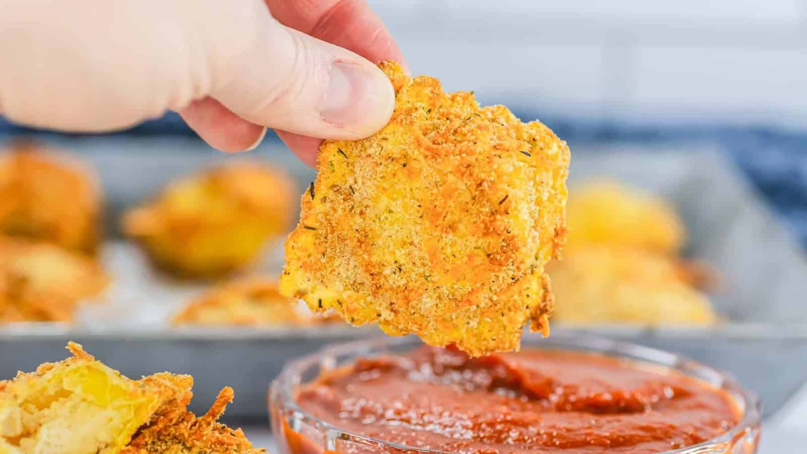 A hand dips a crispy, breaded zucchini chip into a bowl of marinara sauce.