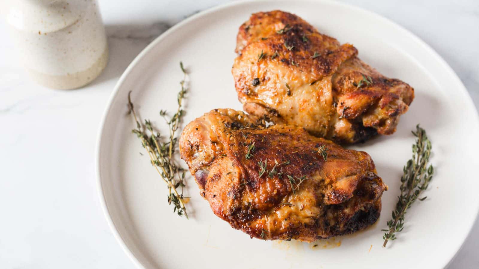 Two roasted chicken thighs on a white plate garnished with sprigs of thyme. A ceramic jar is partially visible in the background on the left.