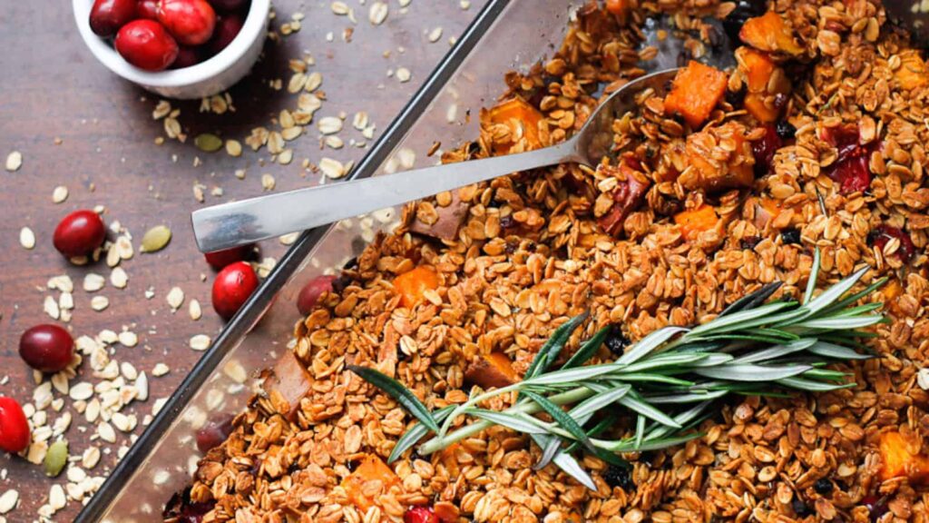 A baking dish filled with a baked oat and fruit mixture, topped with a sprig of rosemary. A spoon rests in the dish. Oats and cranberries are scattered on the table, and a small bowl of cranberries is visible in the background.