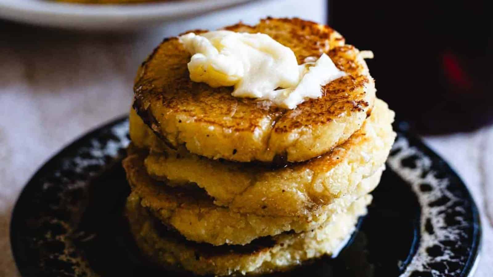 A stack of four golden brown cornmeal pancakes sits on a dark plate, topped with a dollop of melting butter.