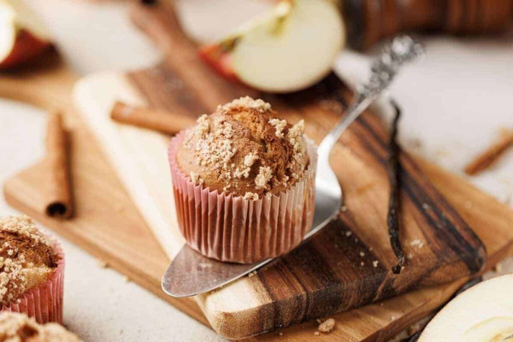 A muffin in a pink paper liner sits on a wooden board with a metal spatula.