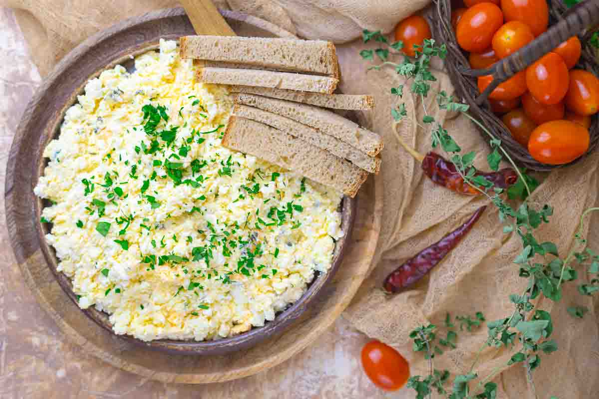 A bowl of egg dip garnished with chopped herbs, served with slices of brown bread, next to cherry tomatoes and fresh herbs on a beige surface.