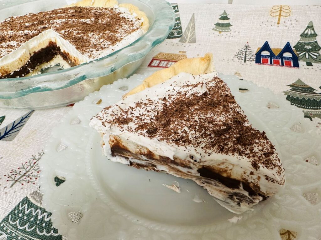 slice of chocolate cream pie, reminiscent of classic black bottom pie, is topped with whipped cream and chocolate shavings on a white plate. The rest of the pie sits in a glass dish on a festive tablecloth with holiday designs.