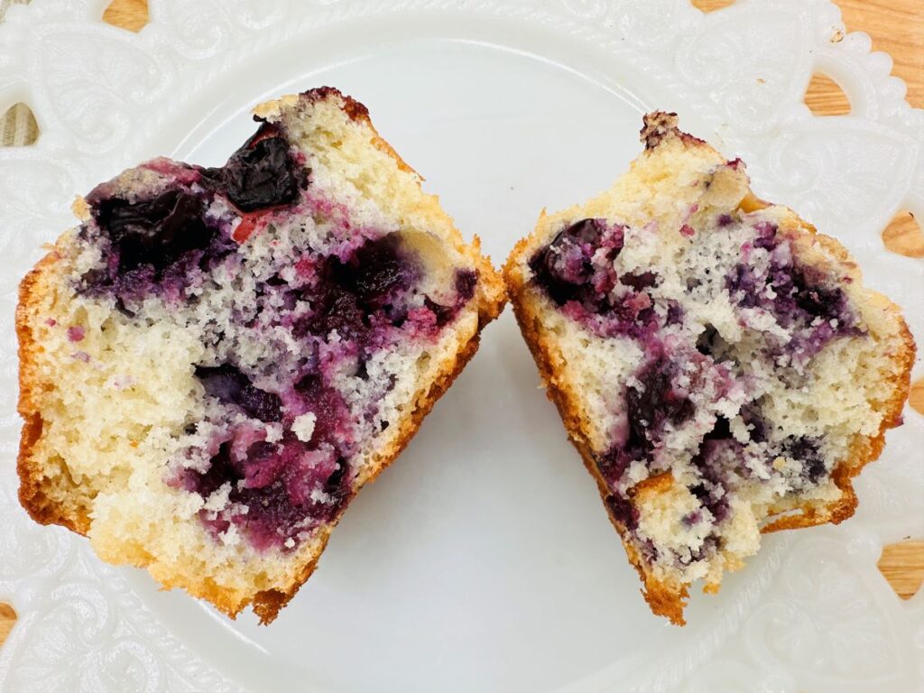 A blueberry muffin cut in half on a white decorative plate, showing a moist interior with visible blueberries.