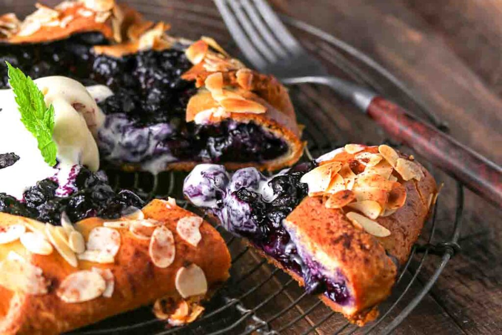 A close-up of a rustic blueberry galette topped with sliced almonds and a dollop of cream, set on a wire rack with a fork nearby.