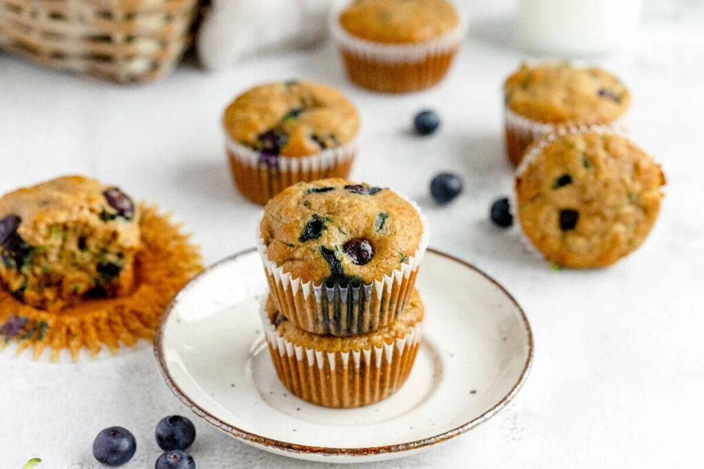 Two blueberry muffins stacked on a plate with scattered blueberries in the background.