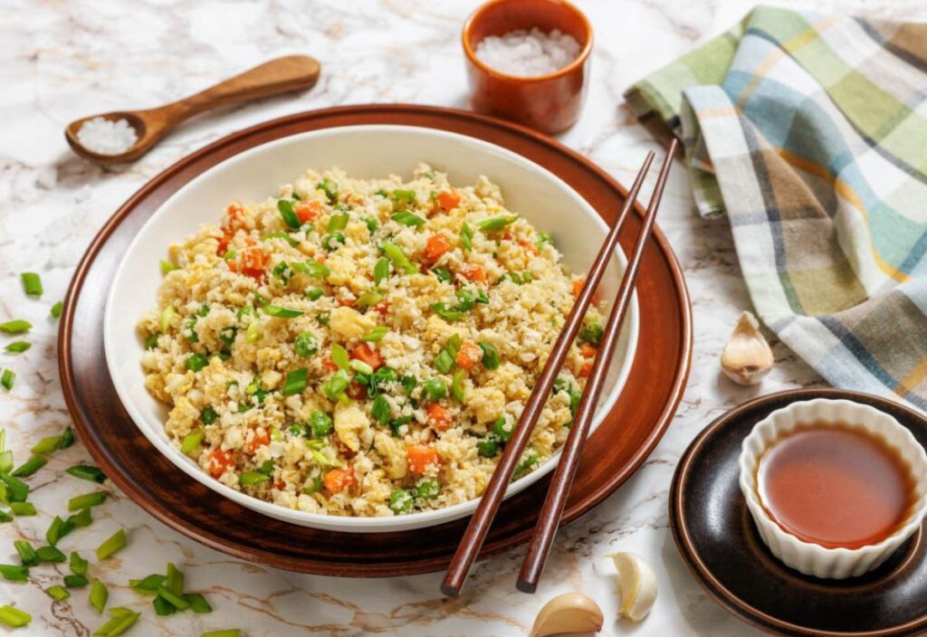 Cauliflower fried rice with vegetables, garnished with green onions, served in a bowl with chopsticks on the side.
