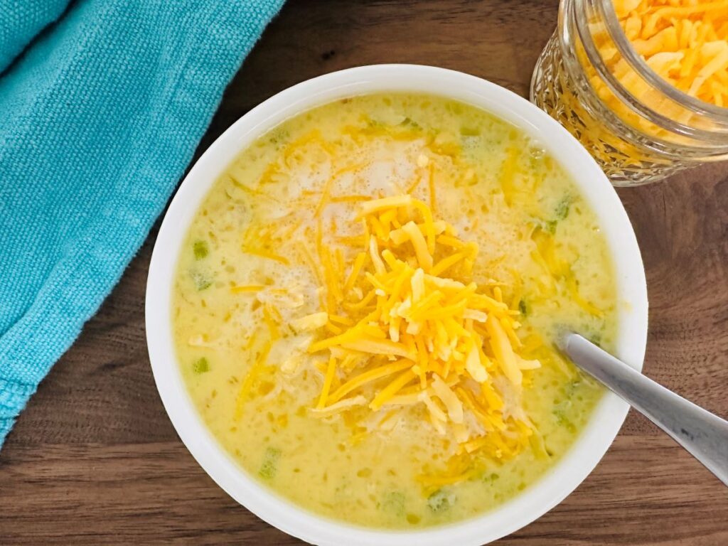 A bowl of creamy cheeseburger chowder topped with shredded cheddar cheese, a spoon inside. A blue cloth sits beside the bowl and a small glass jar filled with more shredded cheese is nearby, all on a wooden surface.