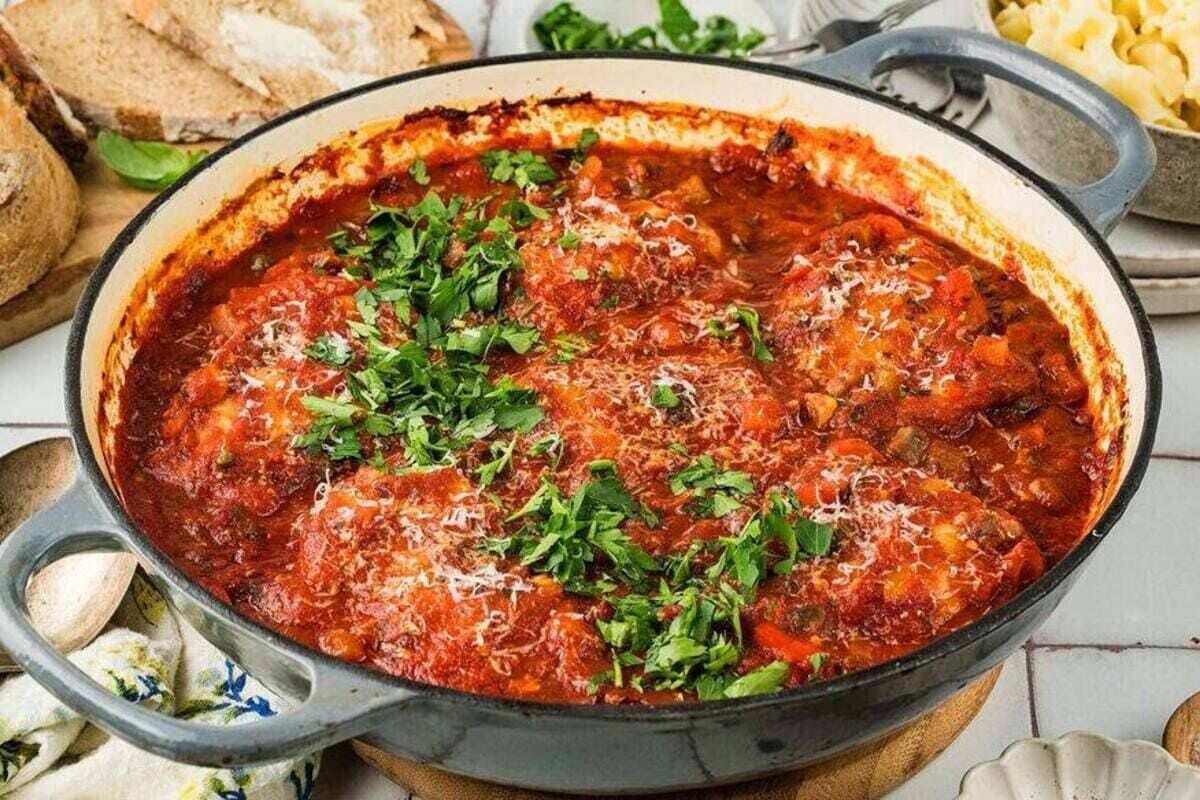 A white dish filled with pieces of Chicken Cacciatore in a red sauce, garnished with chopped herbs.