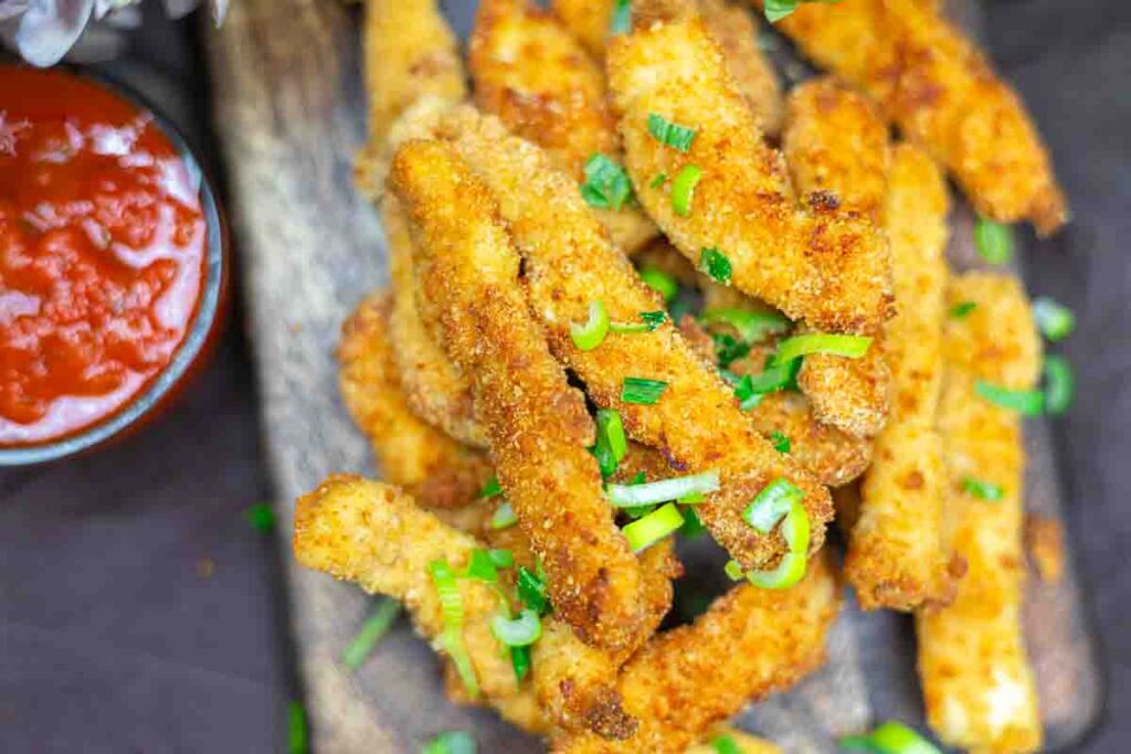 Breaded chicken strips garnished with chopped green onions on a wooden board, with a bowl of red dipping sauce on the side.