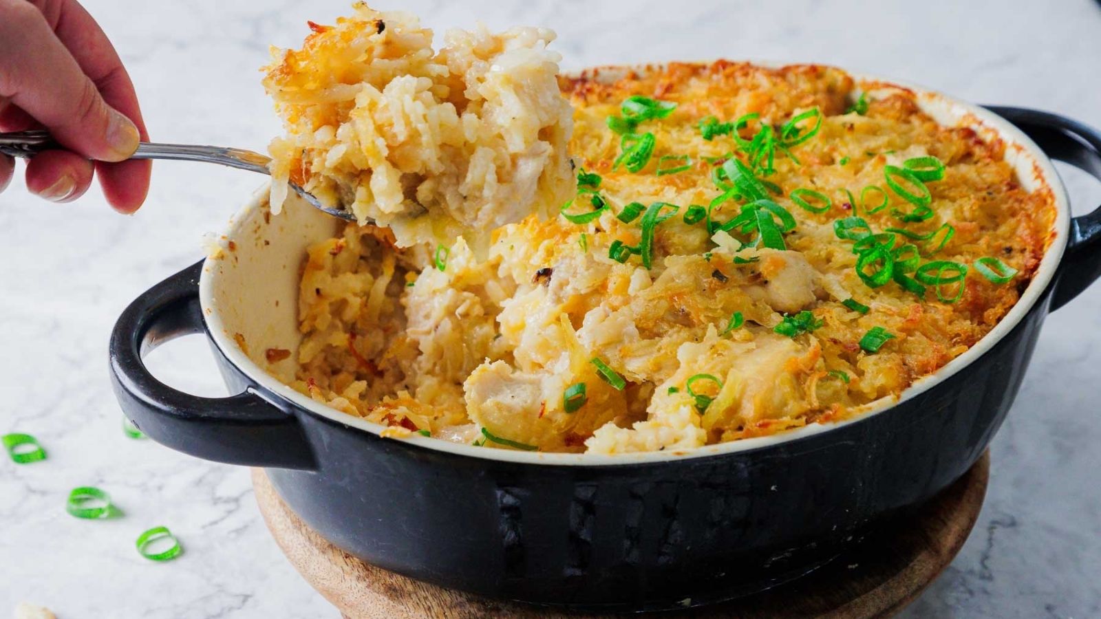 A dish in a black baking pan filled with baked casserole. The top is golden brown with crispy edges and garnished with sliced green onions. A portion has been removed, revealing a creamy interior.