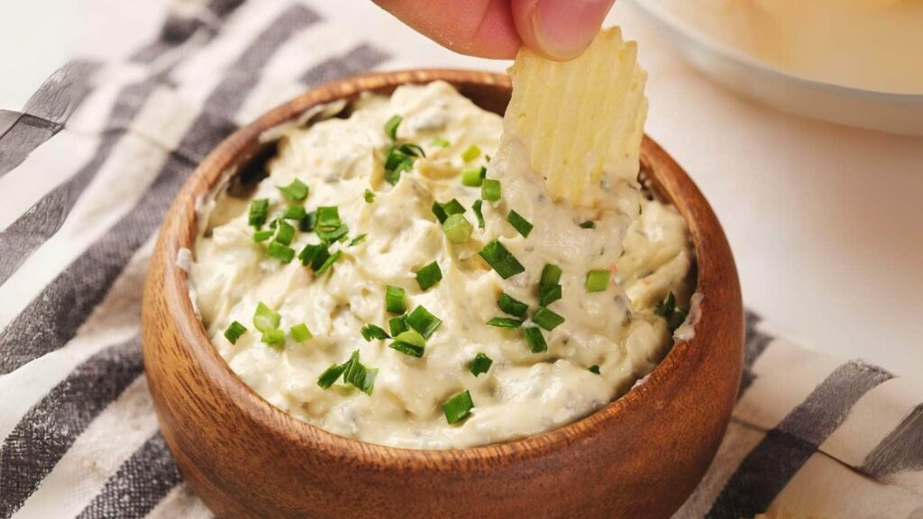 A person's hand dipping a piece of potato chip in a bowl of creamy chip dip.