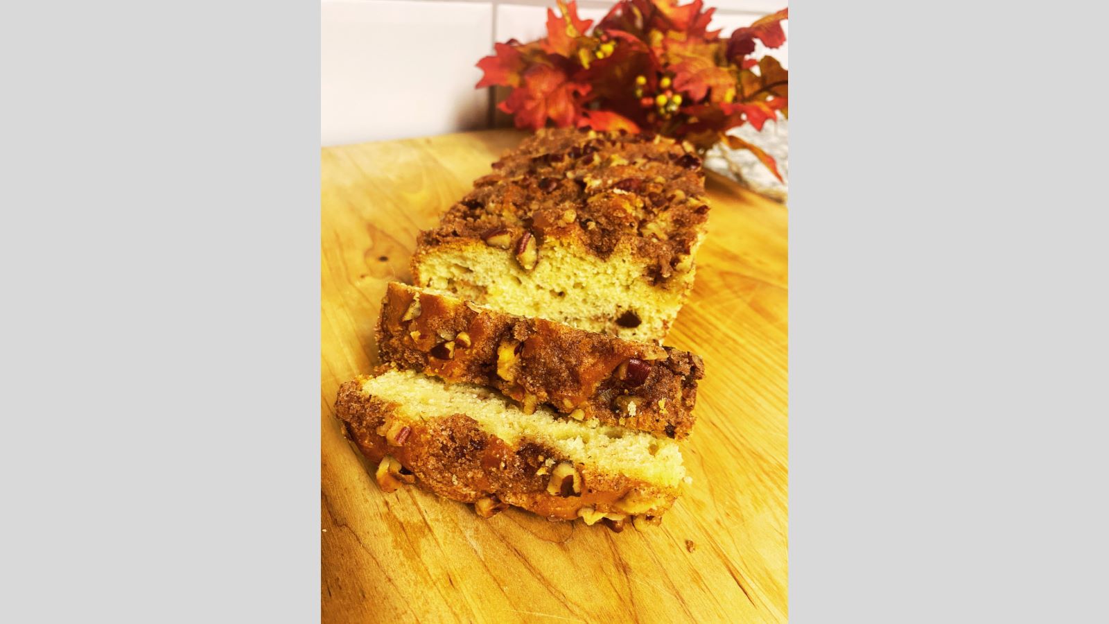 Sliced cinnamon raisin bread on a wooden cutting board, topped with a crumbly sugar and pecan mixture. Autumn leaves are blurred in the background.