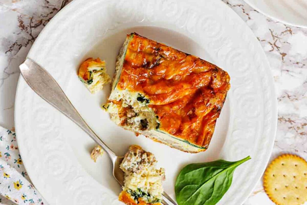 A square slice of baked casserole with visible vegetables and browned cheese on a white plate, with a fork, a spinach leaf, and a cracker nearby.