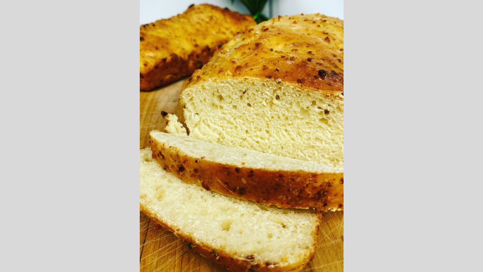 Sliced homemade cottage cheese bread with a golden crust rests on a wooden cutting board. A loaf and blue flowers are in the background, set against a white tiled wall.