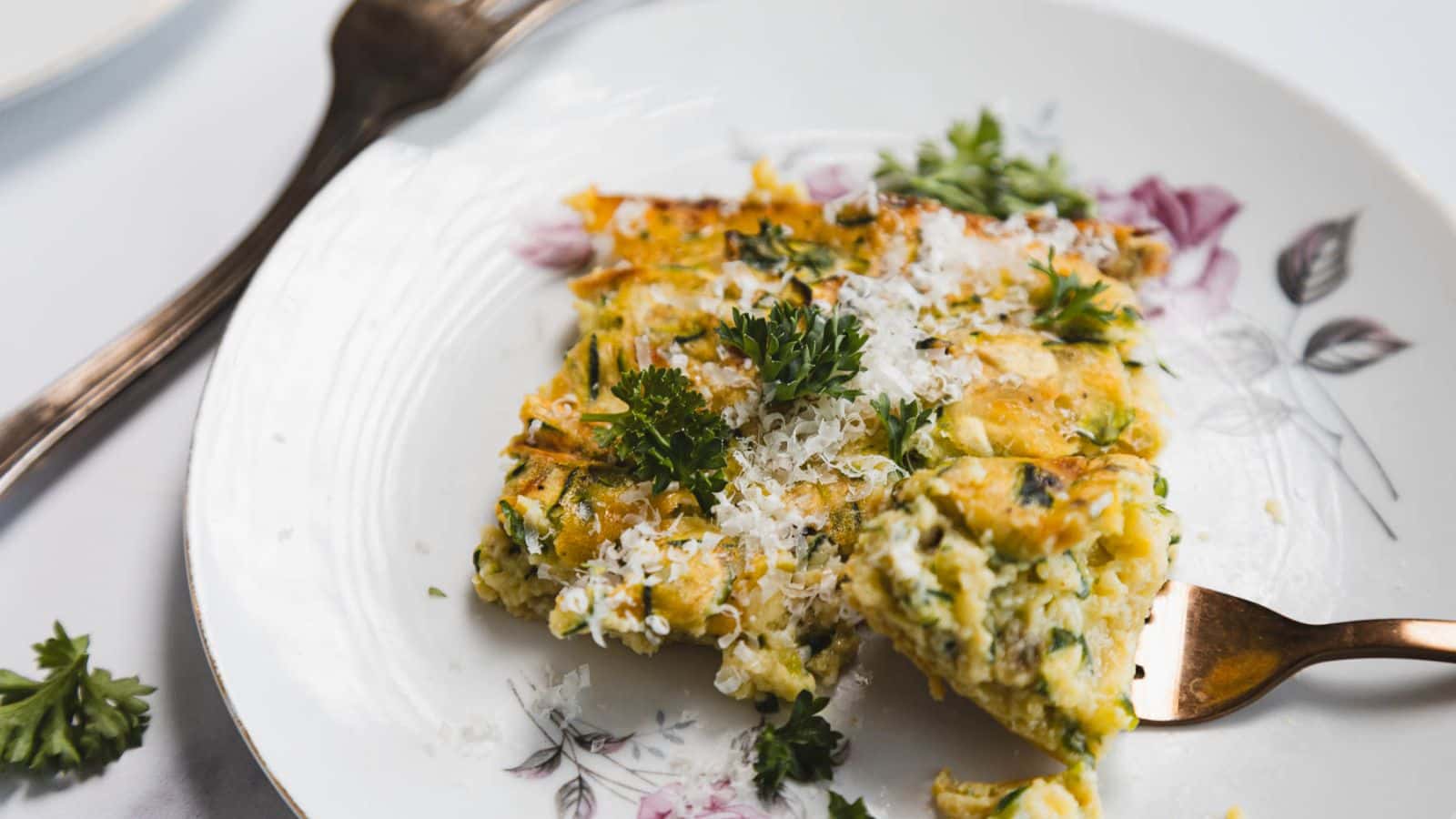 A slice of vegetable frittata topped with grated cheese and parsley sits on a floral plate. A fork is positioned beside the frittata, and another piece of parsley lies on the plate.