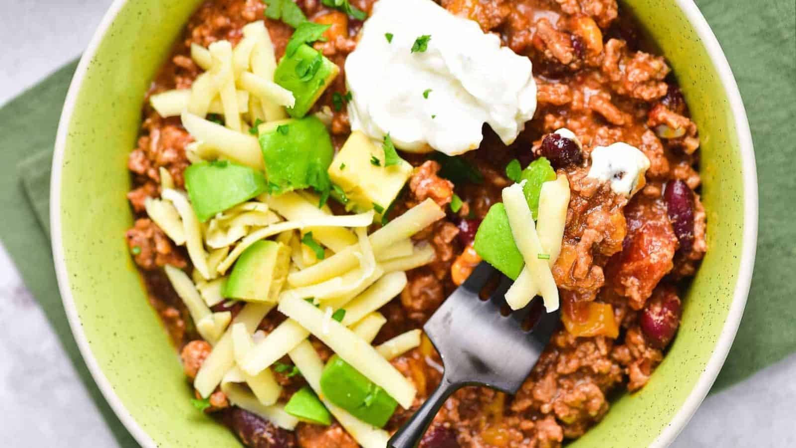 A bowl of chili topped with shredded cheese, diced avocado, and a dollop of sour cream, with a fork resting in the bowl. The food is served in a light green bowl on a green napkin.