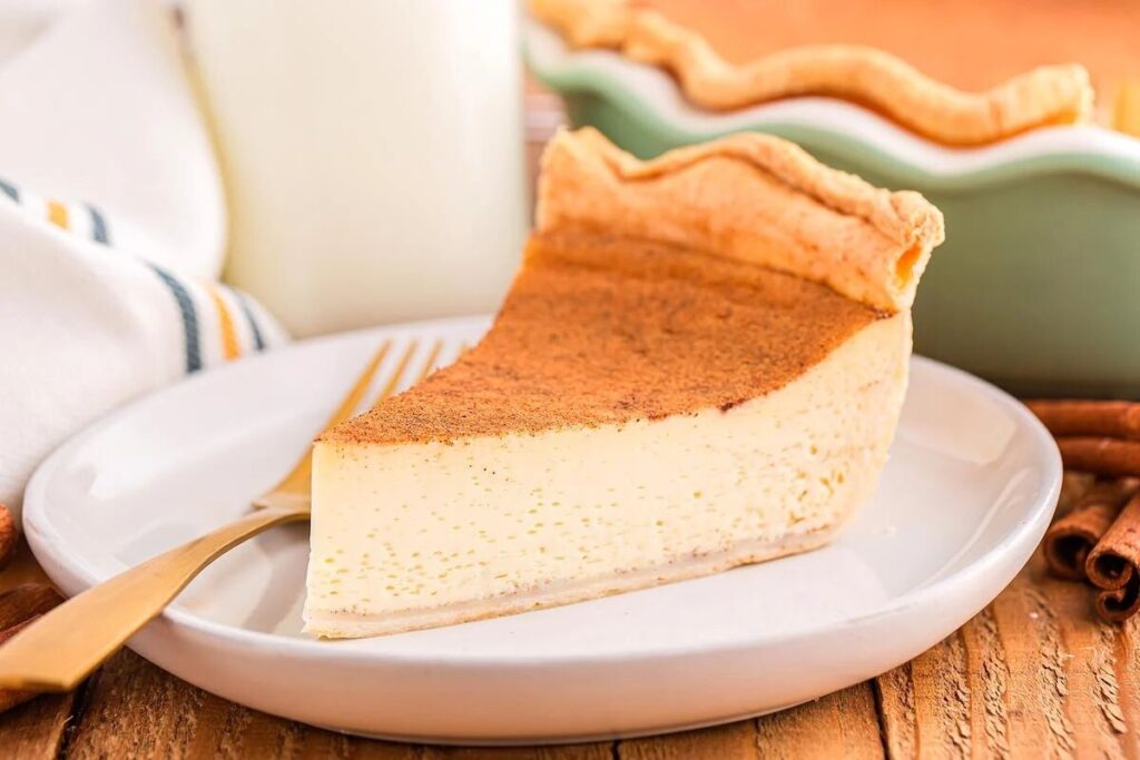 A slice of egg custard pie rests on a white plate with a fork, accompanied by a glass of milk and a pie dish in the background.