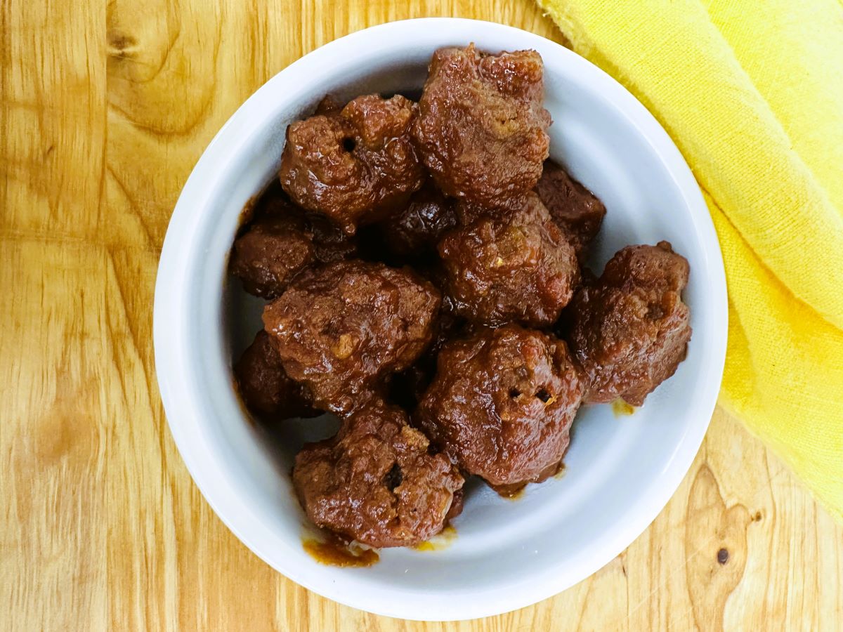 A white bowl filled with several grape jelly and chili sauce meatballs sits on a wooden surface next to a folded yellow cloth.