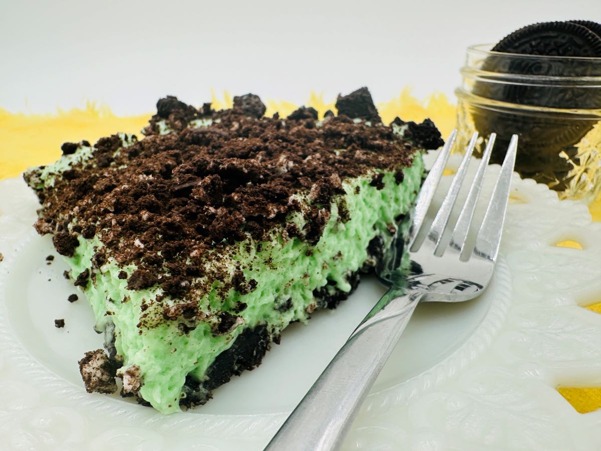 A slice of mint Oreo dessert, reminiscent of grasshopper pie with its green creamy filling and crushed Oreo topping, sits on a white plate with a fork. In the background, a small jar filled with Oreos rests on a yellow surface.