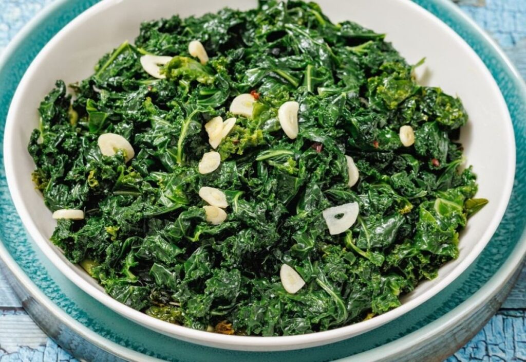 A close-up of a bowl with sautéed kale and garlic slices, served in a white dish with a teal rim.
