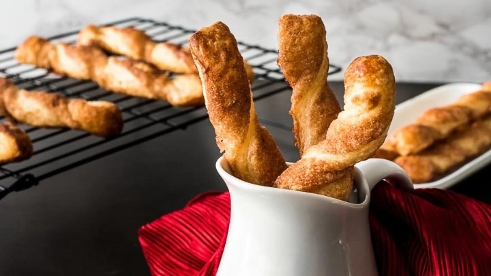 Twisted pastries in a white pitcher with more delicious snacks on a cooling rack and plate in the background. The pitcher is placed on a red fabric, making these kids' treats even more inviting.