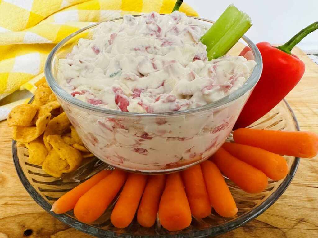 A glass bowl of creamy Hot Chipped Beef Dip with chopped red pieces, garnished with a celery stick. Baby carrots, corn chips, and a whole red pepper surround the bowl on a glass plate. A yellow checked cloth is in the background.