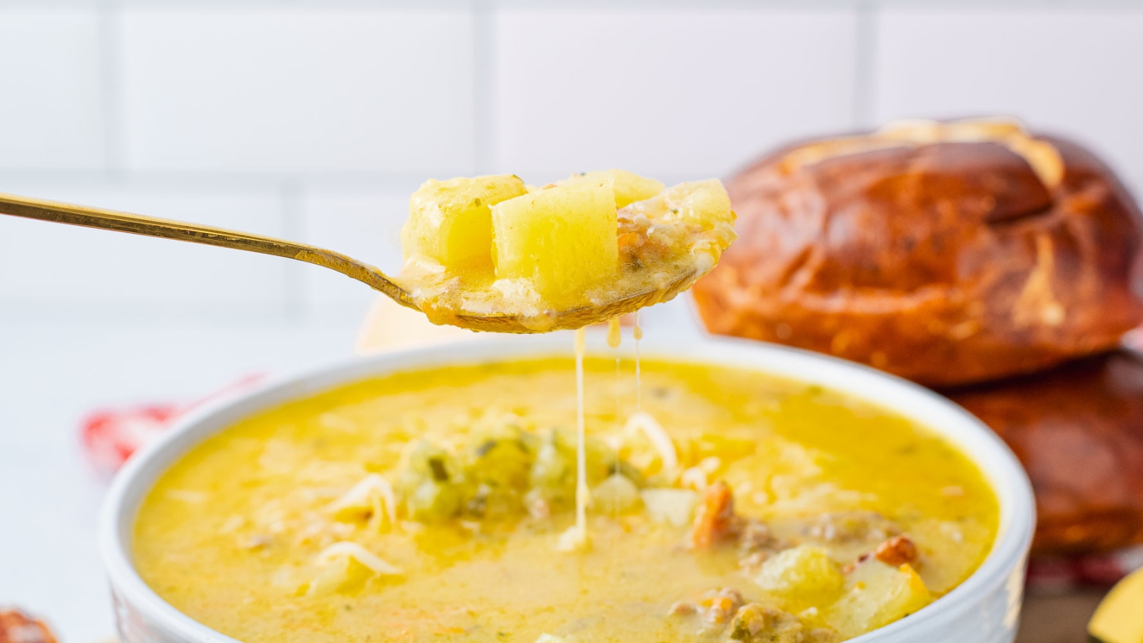 A bowl of cheesy soup with ground meat, potatoes, and vegetables, served with a pretzel bun.