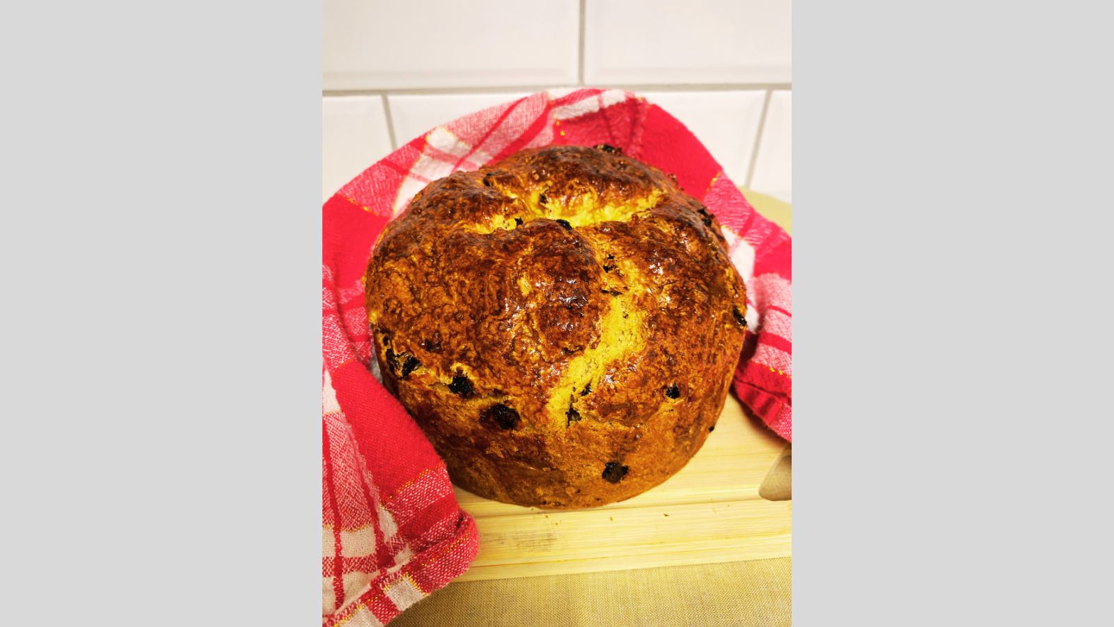 A round loaf of golden-brown Irish Soda Bread with raisins cools on a wire rack. The bread has a shiny, crisp crust and rests on a speckled countertop with white tile in the background.