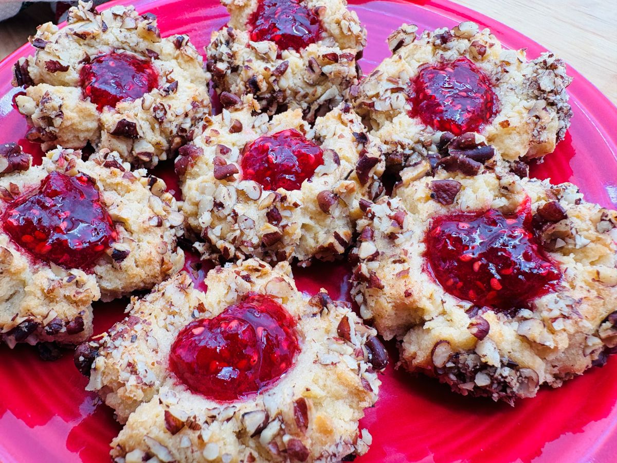 Close-up of thumbprint cookies topped with chopped nuts and filled with red jam, arranged on a red surface.
