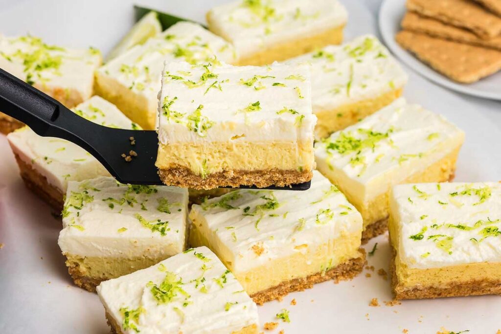 A spatula lifts a creamy key lime bar with a graham cracker crust, topped with fresh lime zest.