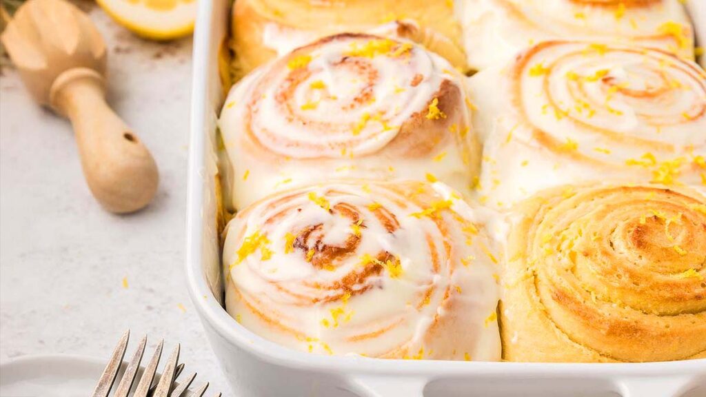A baking dish with frosted cinnamon rolls topped with lemon zest, surrounded by a wooden utensil and a silver fork.